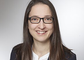 Porträtfoto einer lächelnden Frau mit dunkler Brille und langen braunen Haaren. Sie trägt eine weiße Bluse und ein dunkles Sakko. Neutraler Hintergrund, professionelle Aufnahmesituation.