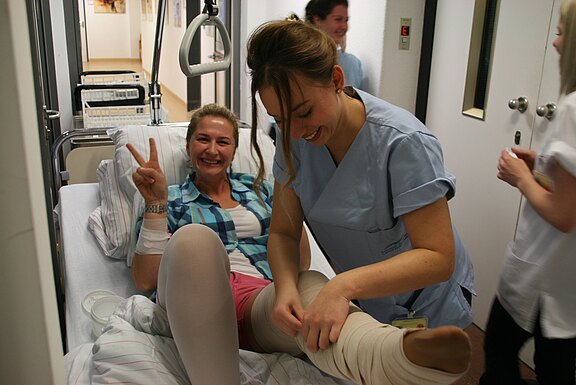Patientin liegt lächelnd im Krankenhausbett und zeigt das Victory-Zeichen, während eine Pflegekraft ihr am Bein einen Verband anlegt. Die Szene vermittelt medizinische Versorgung und eine positive, unterstützende Stimmung.
