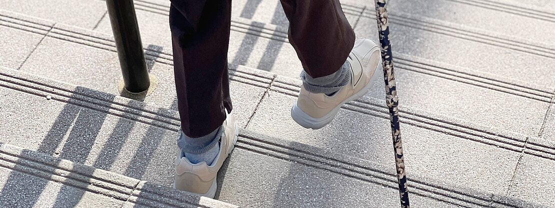 Eine Person mit einer weißen Jacke und dunklen Hosen geht eine Treppe hinunter, während sie sich mit einem blumigen Gehstock abstützt. Ihre Schuhe sind hell und sie hat graue Socken an. Die Stufen sind mit Rillen versehen.
