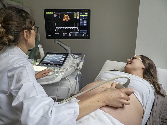 Eine schwangere Frau liegt auf einer Liege und lächelt, während eine Ärztin ein Ultraschallgerät bedient. Der Fokus liegt auf dem Bauch der Frau, wobei ein Ultraschallbild auf dem Bildschirm zu sehen ist. Die Atmosphäre ist beruhigend und informativ.