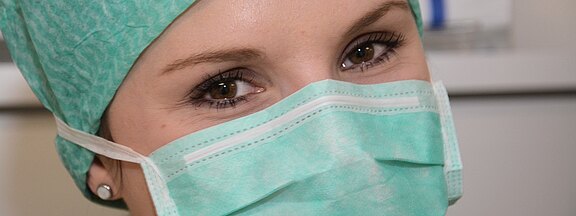 Nahaufnahme einer medizinischen Fachkraft mit grüner OP-Mütze und chirurgischer Maske; nur die Augen sind sichtbar. Aufmerksamer Blick, Symbol für Hygiene und Gesundheitsschutz.