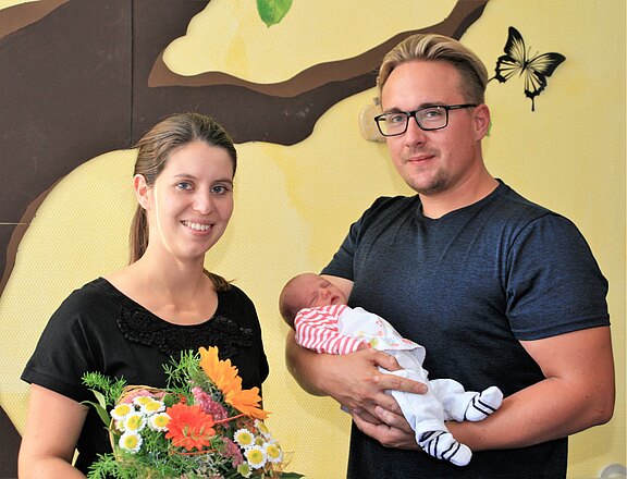Ein Paar steht vor einer hellen Wand mit einem Baummotiv. Die Frau hält einen Strauß bunter Blumen, während der Mann ein neugeborenes Baby im Arm trägt. Beide lächeln glücklich in die Kamera.