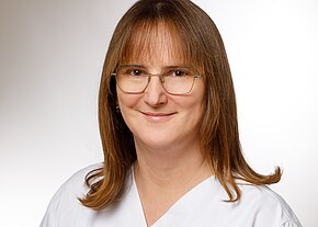 Porträt einer lächelnden erwachsenen Person mit schulterlangem braunem Haar und Brille, frontal vor neutralem Hintergrund. Sie trägt weiße Arbeitskleidung.