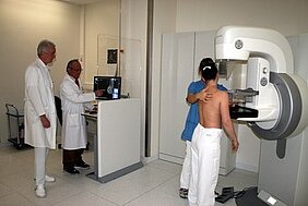 Ein Kind steht vor einem medizinischen Gerät zur Untersuchung, während ein Arzt daneben am Computer arbeitet. Eine Person in blauer Kleidung assistiert dem Kind, das sich für die Untersuchung vorbereitet. Im Hintergrund sind weitere Fachleute zu sehen.