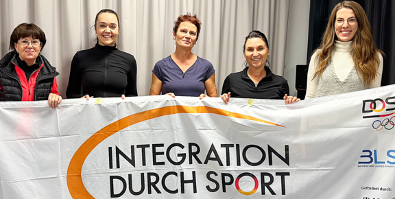 Fünf Frauen halten gemeinsam ein großes Banner mit der Aufschrift „Integration durch Sport“. Sie stehen nebeneinander und lächeln; die Botschaft betont Zusammenhalt und Teilhabe durch Sport.