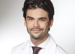 Dr. med. Marcel Hoh Porträt eines jungen Mannes mit dunklem Haar und kurzem Bart. Er trägt weißen Arztkittel und Krawatte, lächelt leicht, vor neutralem Hintergrund. Professionelles, seriöses Profilfoto.