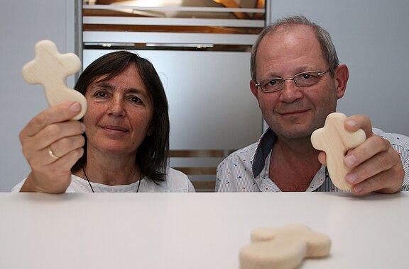 Zwei Personen blicken in die Kamera und halten jeweils ein kleines, helles Holzkreuz hoch; ein weiteres Holzkreuz liegt im Vordergrund.