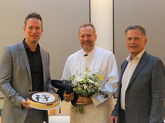 Drei Männer posieren drinnen; der mittlere trägt weißen Arztkittel und hält einen Blumenstrauß. Der linke Mann überreicht eine Torte mit einfachem Figurenmotiv. Der rechte steht daneben. Alle lächeln; der Anlass wirkt wie Gratulation oder Ehrung.