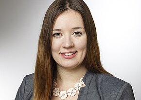 Portrait einer lächelnden Frau mit langen, braunen Haaren und einem grauen Blazer. Sie trägt eine auffällige Halskette mit blütenförmigen Anhängern. Der Hintergrund ist hell und einfarbig, was den Fokus auf ihr Gesicht lenkt.