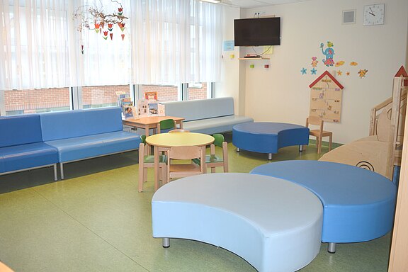 Heller Kinderbereich zum Warten und Spielen mit blauen Sofas, geschwungenen Sitzinseln, zwei runden Tischen und kleinen Stühlen. Große Fenster mit Vorhängen; an der Wand Deko für Kinder und ein Fernseher. Freundliche, aufgeräumte Atmosphäre.