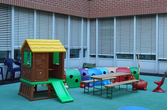 Kinderspielbereich im Innenhof: kleines Kunststoff-Spielhaus mit gelbem Dach und grüner Rutsche, daneben ein bunter Kriechtunnel in Raupenform. Davor stehen Tische und Bänke auf weichem, türkisfarbenem Boden.