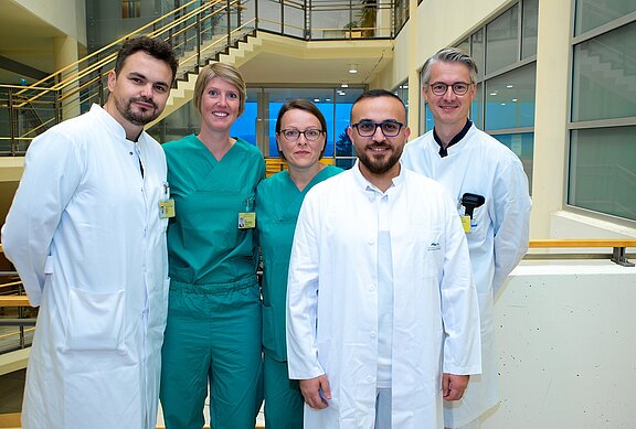 Fünf medizinische Fachkräfte stehen nebeneinander in einem hellen Krankenhausbereich. Drei tragen weiße Kittel, zwei grüne OP-Kleidung. Alle schauen in die Kamera, freundlich und professionell. Klinikumgebung im Hintergrund.