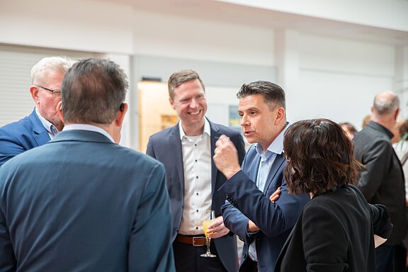 Mehrere Erwachsene in Businesskleidung unterhalten sich lebhaft bei einem Empfang. Ein Mann gestikuliert mit einem Getränk in der Hand, andere hören lächelnd zu. Atmosphäre: berufliches Netzwerkgespräch.