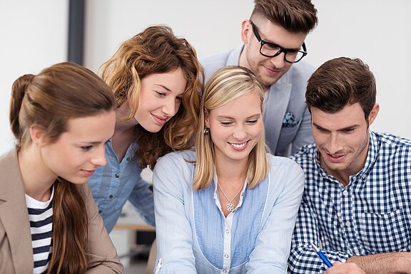 Sechs Personen stehen um einen Tisch und schauen konzentriert auf etwas, das sich dort befindet. Sie scheinen an einem Projekt oder einer Diskussion zu arbeiten, wobei ihre Mienen interessiert und engagiert wirken.