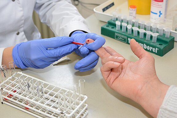 Eine Hand mit blauen Handschuhen führt eine Blutentnahme an einer hellhäutigen Hand durch. Auf dem Tisch sind Reagenzgläser und andere Labormaterialien sichtbar, die für die Analyse verwendet werden.