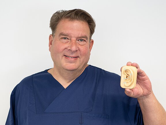 Mann in blauer OP-Kleidung hält lächelnd ein Ohrmodell vor hellem Hintergrund, als würde er eine HNO- oder Hörberatung darstellen.