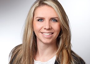 Nahaufnahme eines Porträtfotos: lächelnde junge Frau mit langen blonden Haaren, blickt in die Kamera. Heller, neutraler Hintergrund; Business-Kleidung, professionelles Bewerbungsfoto.