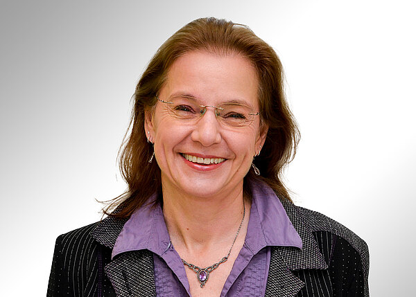 Porträt einer lächelnden Frau mit Brille, mittellangem braunem Haar, lila Bluse mit Halskette und dunklem Blazer vor neutralem Hintergrund.