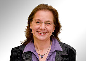 Cécile Koubek Porträt einer lächelnden Frau mit Brille, mittellangem braunem Haar, lila Bluse mit Halskette und dunklem Blazer vor neutralem Hintergrund.