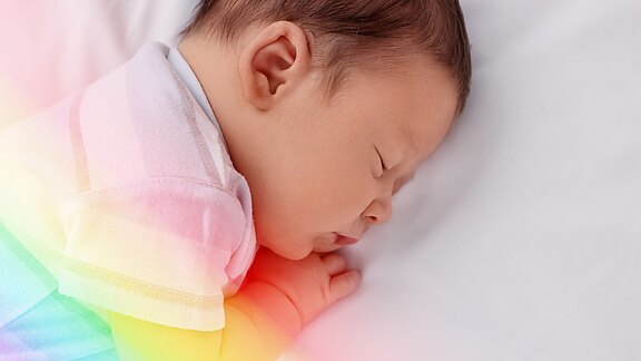 Schlafendes Baby auf der Seite auf weißer Bettwäsche, Gesicht entspannt, Hand unter der Wange. Weiches, buntes Licht in Regenbogenfarben fällt über den Körper.
