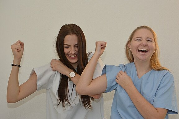 Zwei Frauen in Ärztekitteln lachen und freuen sich miteinander. Sie heben ihre Arme in einer positiven Geste, während sie sich an ihren Oberarmen berühren. Der Hintergrund ist neutral.