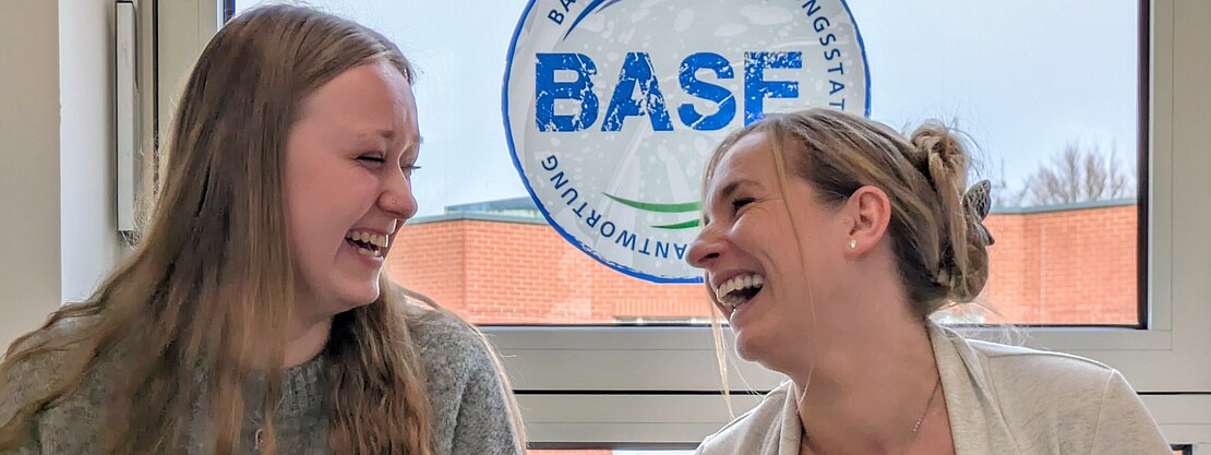 Zwei junge Frauen sitzen nebeneinander in einem Raum und lachen sich herzlich an. Tageslicht fällt durch ein Fenster. Die Szene vermittelt eine lockere, freundliche Stimmung wie bei einer entspannten Unterhaltung.