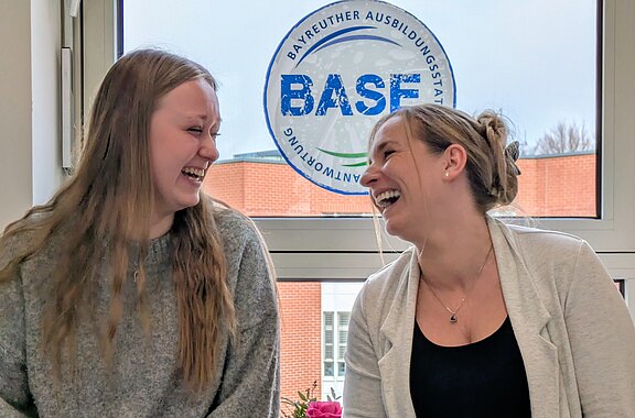 Zwei junge Frauen sitzen nebeneinander in einem Raum und lachen sich herzlich an. Tageslicht fällt durch ein Fenster. Die Szene vermittelt eine lockere, freundliche Stimmung wie bei einer entspannten Unterhaltung.