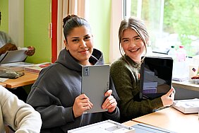 Zwei Jugendliche sitzen an einem Tisch und lächeln vor der Kamera. Sie halten jeweils ein Tablet in den Händen. Im Hintergrund sind weitere Schüler und einige Gegenstände auf dem Tisch zu sehen. Die Atmosphäre wirkt freundlich und aktiv.