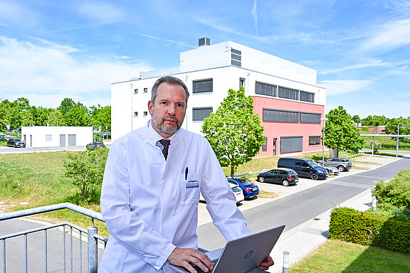 Person im weißen Kittel arbeitet an einem Laptop auf einer Terrasse; im Hintergrund modernes Klinik- oder Forschungsgebäude und Parkplatz an einem sonnigen Tag.