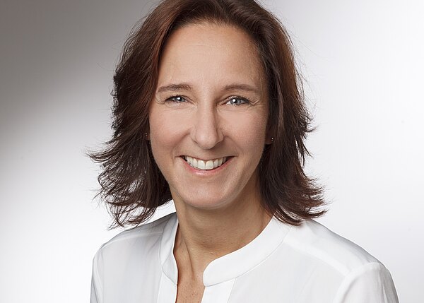 Porträtfoto: Eine lächelnde Frau mit schulterlangem braunem Haar blickt in die Kamera. Sie trägt eine weiße Bluse vor neutralem, hellgrauem Hintergrund; freundlicher, professioneller Ausdruck.