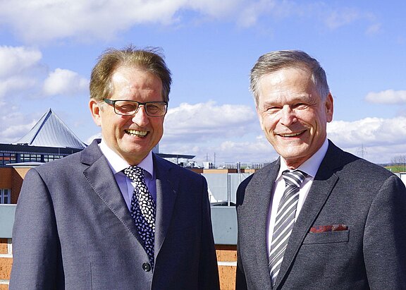 Zwei Männer mittleren bis höheren Alters in Anzügen stehen nebeneinander und lächeln in die Kamera. Heller Sonnenschein, blauer Himmel. Hintergrund unscharfe Gebäude; Fokus auf freundliches, professionelles Porträt im Freien.