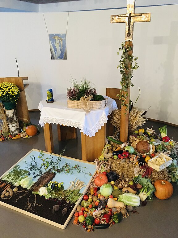Kirchlicher Altar mit großem Kreuz und Erntedankgaben: Körbe, Getreide, Brot, viele Obst‑ und Gemüsesorten sowie Kürbisse auf Stroh. Auf dem Boden ein Bild eines Baums aus Erde und Naturmaterialien. Festliche Dekoration, die Dank für die Ernte ausdrückt.