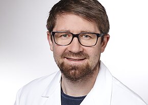 Porträt eines lächelnden Mannes mit Brille, braunem Kurzhaar und Bart. Er trägt einen weißen Laborkittel über dunklem Shirt. Heller, neutraler Hintergrund.