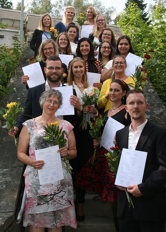Eine Gruppe von 17 Personen steht auf einer Treppe, hält Blumen und Urkunden in den Händen. Sie lächeln und feiern offenbar einen Abschluss oder eine Ehrung. Der Hintergrund zeigt Begrünung und teilweise einen klaren Himmel.