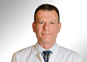 Porträt eines Mannes in weißem Arztkittel und Krawatte, frontal aufgenommen vor neutralem Hintergrund, mit freundlichem, professionellem Gesichtsausdruck.