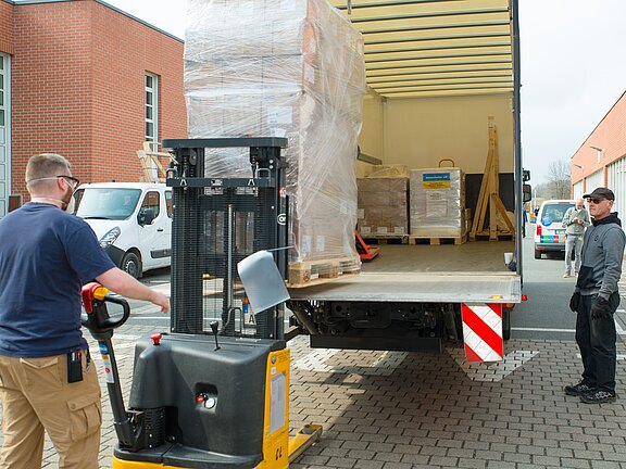 Zwei Lagerarbeiter laden mit einem elektrischen Hubwagen eine in Folie verpackte Palette in den geöffneten Laderaum eines Lieferwagens. Weitere Paletten stehen bereits im Fahrzeug. Szene eines Warenumschlags auf dem Hof.