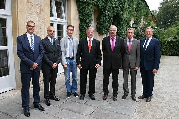 Sieben Männer stehen nebeneinander für ein Gruppenfoto vor einem historischen Gebäude mit Efeu. Die meisten tragen Anzüge; einer ist leger mit Jeans und Hemd. Formelles, professionelles Treffen im Freien.