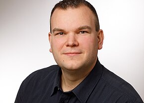 Porträtfoto einer Person mit kurzem dunklem Haar und leichtem Lächeln, die direkt in die Kamera blickt. Sie trägt ein dunkelgraues Hemd; der Hintergrund ist hell und neutral, wie bei einem professionellen Bewerbungsfoto.