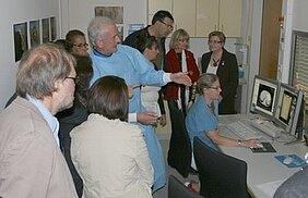 Eine Gruppe von Menschen steht um einen Monitor, auf dem medizinische Bilder zu sehen sind. Eine Person im Vordergrund erklärt die Bilder, während die anderen aufmerksam zuhören. Die Atmosphäre ist von Interesse und Austausch geprägt.