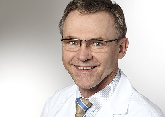 Porträt eines lächelnden Mannes mittleren Alters mit Brille im weißen Arztkittel, Hemd und gestreifter Krawatte vor neutralem Hintergrund.