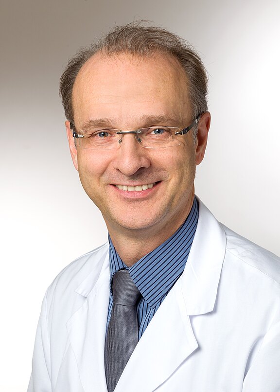 Porträt eines lächelnden Mannes mittleren Alters mit Brille in weißem Arztkittel, blau gestreiftem Hemd und grauer Krawatte vor neutralem Hintergrund.