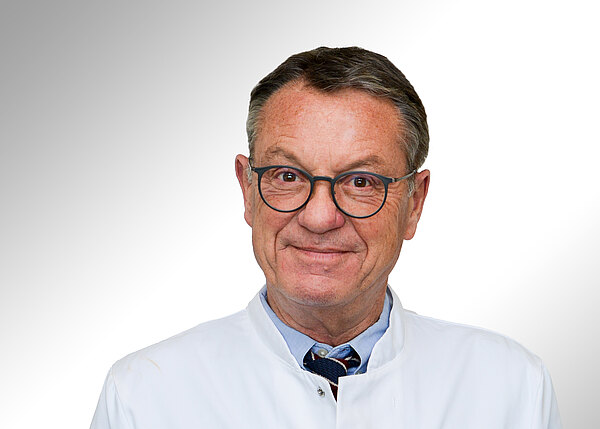 Porträt eines älteren Mannes im weißen Arztkittel mit runder Brille. Er lächelt freundlich und blickt direkt in die Kamera vor hellem, neutralem Hintergrund.