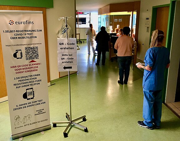 Innenraum eines Testzentrums: Aufsteller mit Anweisungen zur Selbstregistrierung für einen COVID‑19‑Test und QR‑Code. Einige Personen warten vor einem Schalter; eine Pflegekraft in blauer Kleidung hält ein Klemmbrett. Atmosphäre einer organisierten Anmeldung zu Coronatests.