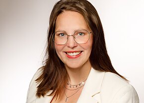 Porträtfoto: lächelnde Person mit mittellangen braunen Haaren und silberner Brille, trägt weißen Blazer und Ketten, vor neutral hellem Hintergrund.