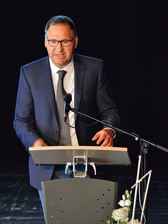 Mann im Anzug spricht am Rednerpult auf einer Bühne; Mikrofon vor ihm. Er gestikuliert und blickt nach vorn. Die Szene wirkt wie eine formelle Rede oder Präsentation.