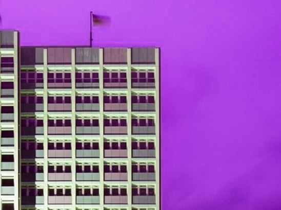 Modernes Hochhaus mit rasterförmiger Fassade und vielen identischen Fenstern vor einem intensiven violetten Himmel; auf dem Dach weht eine kleine Fahne.