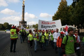 Eine Gruppe von Menschen in leuchtenden T-Shirts marschiert auf einer Straße und trägt ein großes Banner mit der Aufschrift "Klinikum Bayreuth". Im Hintergrund ist ein Monument zu sehen. Die Atmosphäre wirkt demonstrativ und engagiert.