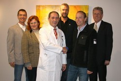 Sechs Personen stehen in einer Gruppe vor einem Kunstwerk. Ein Mann in einem weißen Arztkittel schüttelt einem anderen Mann die Hand. Die anderen Personen beobachten diesen Moment. Die Szene vermittelt ein Gefühl von Professionalität und Zusammenarbeit.