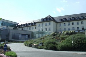 Eine moderne Gebäudestruktur mit einem zweigeschossigen Teil, geschmückt mit Dachverzierungen und großen Fenstern. Vor dem Gebäude erstreckt sich eine gepflegte Grünfläche mit Sträuchern. Es ist ein heller, sonniger Tag.