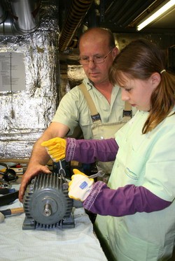 Ein Erwachsener und ein Kind arbeiten gemeinsam an einem Elektromotor. Der Erwachsene zeigt dem Kind, wie man mit Werkzeugen umgeht. Beide tragen Schutzhandschuhe und konzentrieren sich auf ihre Aufgabe in einer Werkstatt.